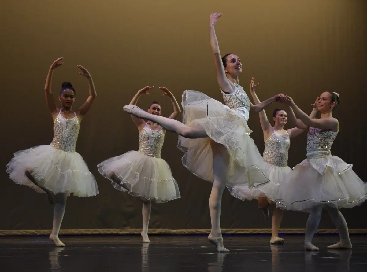 Teen ballet dancers in white tutus perform on stage in arabesque