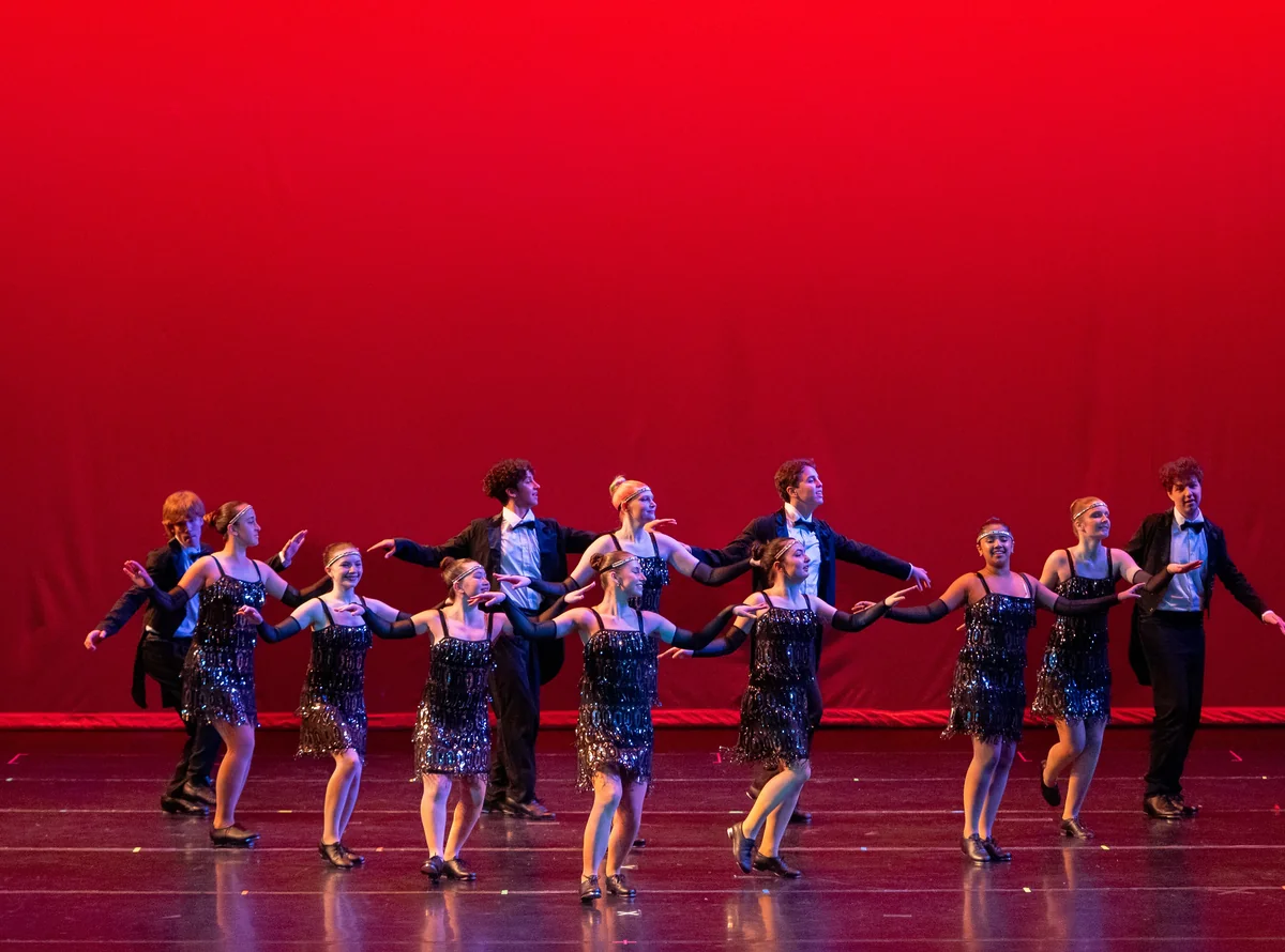 Tap dancers in flapper costumes and tuxedos perform on stage in tap shoes