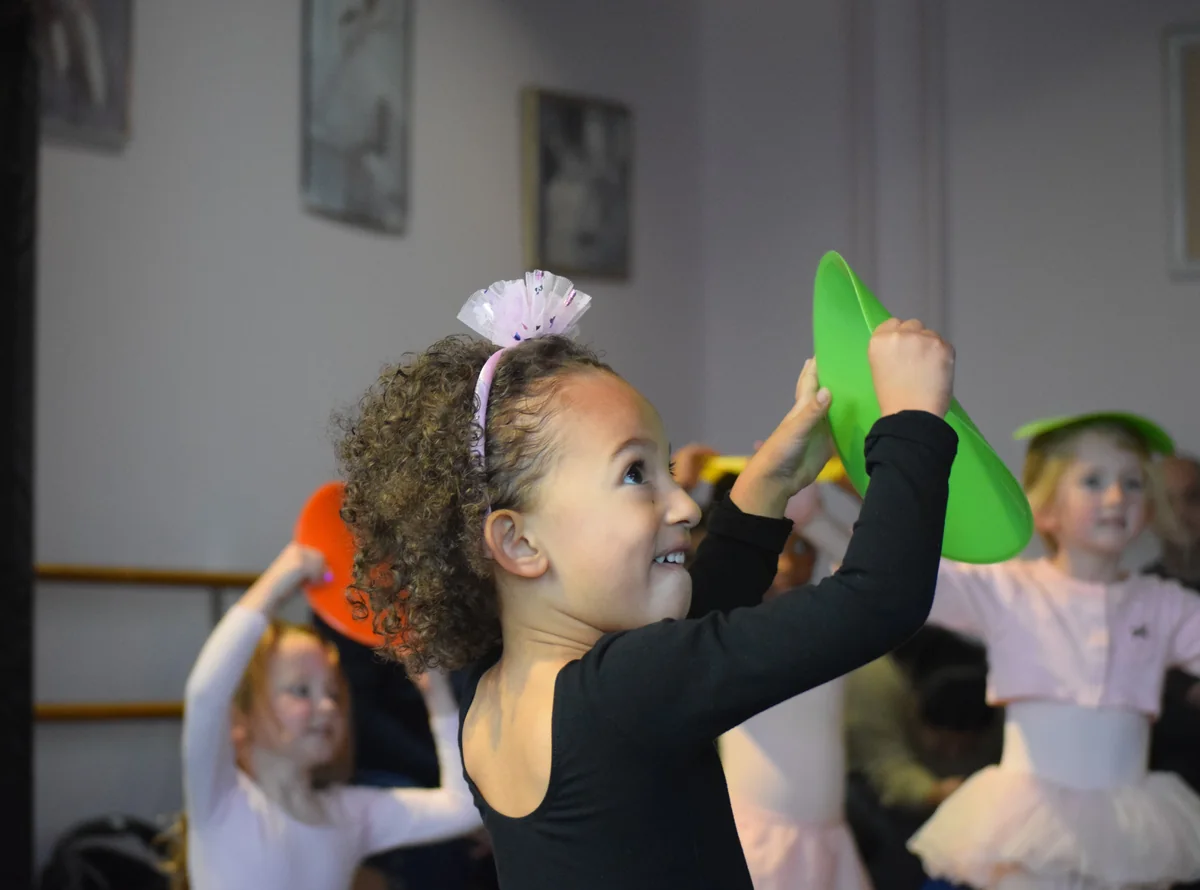 Young dancer smiles as she lifts a green disc overhead during a Turning Tots class
