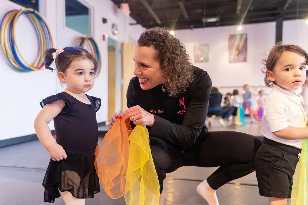 A toddler in a black leotard holds a colourful scarf beside her teacher during a Grow With Us dance class for ages 2 to 7